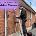 Technician on a ladder using a laptop to configure audio on a Hikvision security camera.